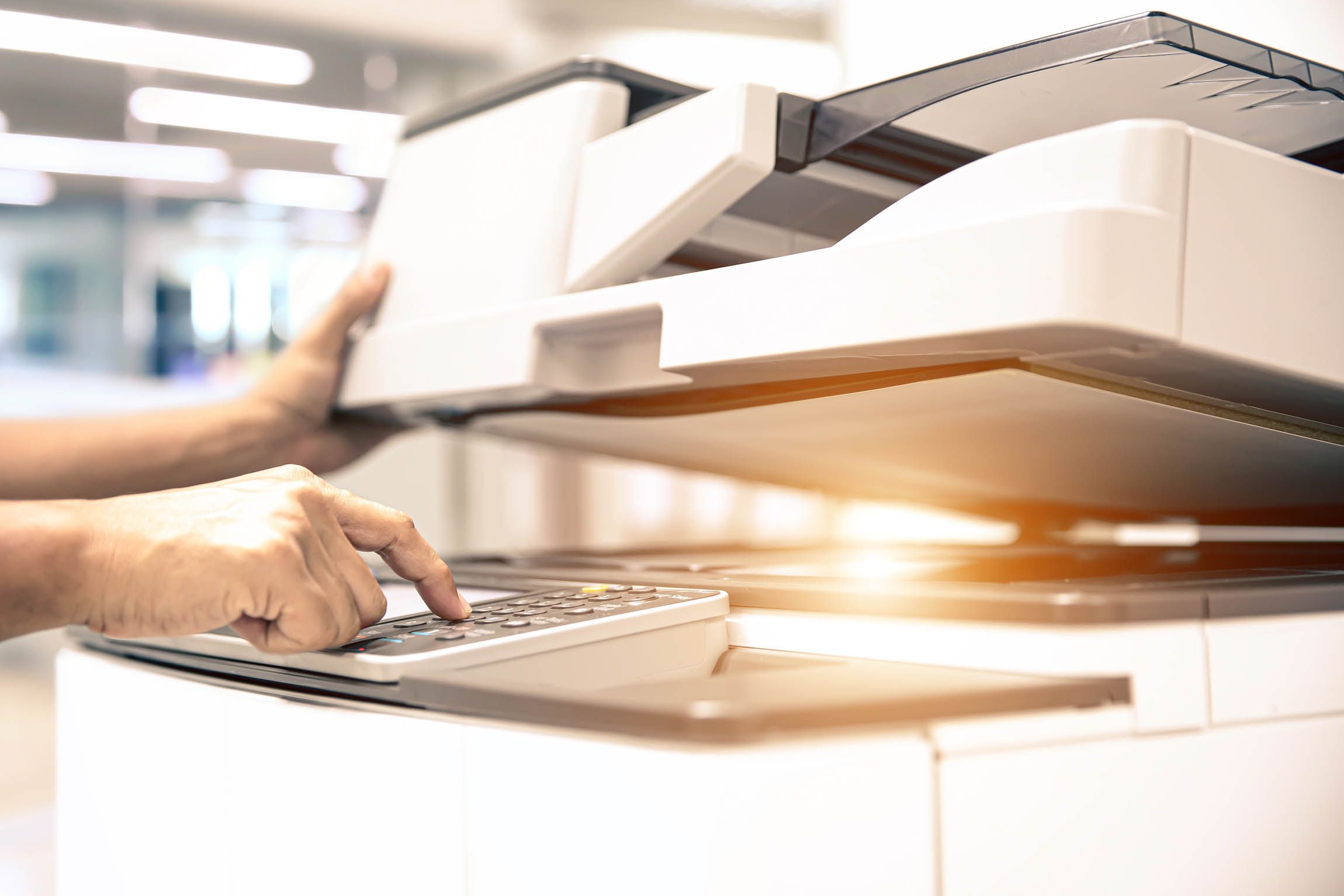 Hand bedient das Bedienfeld eines weißen Kopiergeräts in einem Büro mit unscharfem Hintergrund.